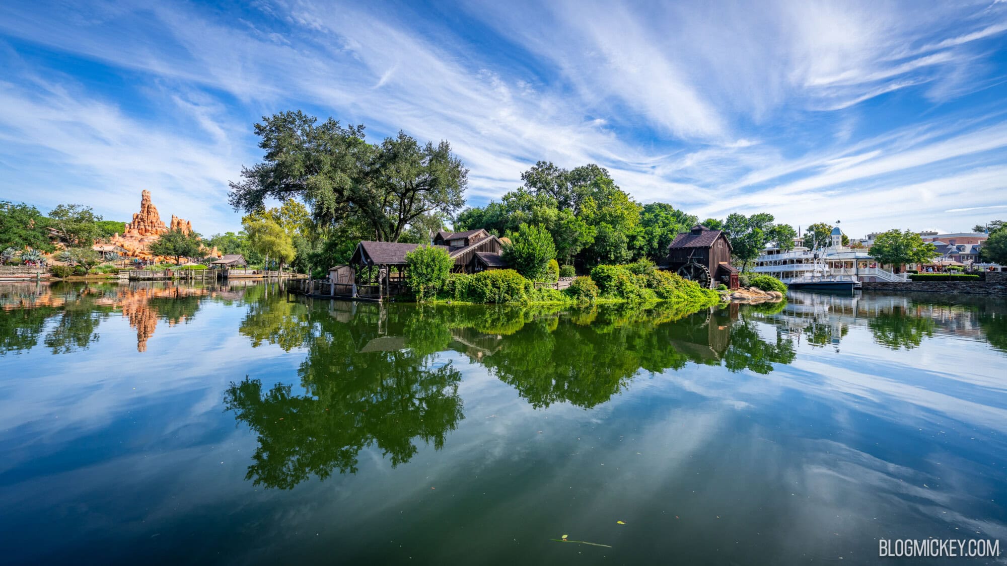 Lush green trees and rustic buildings surrounding a calm Rivers of America with clear reflections at Walt Disney World's Frontierland, featuring the Big Thunder Mountain Railroad mountain in the background and lush Tom Sawyer Island featured
