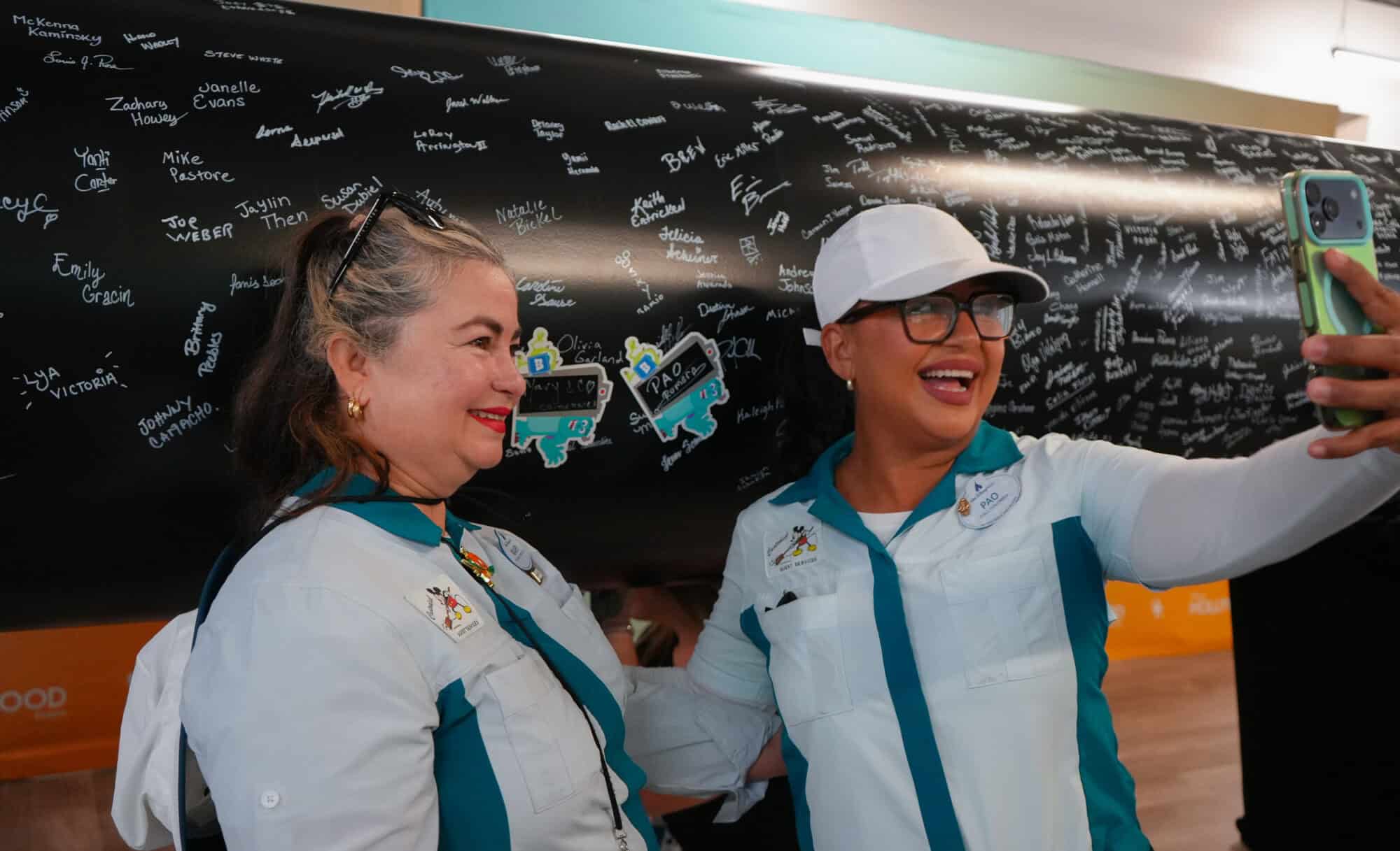 Two cast members pose for a selfie in front of a signed wall at Monstropolis milestone event.