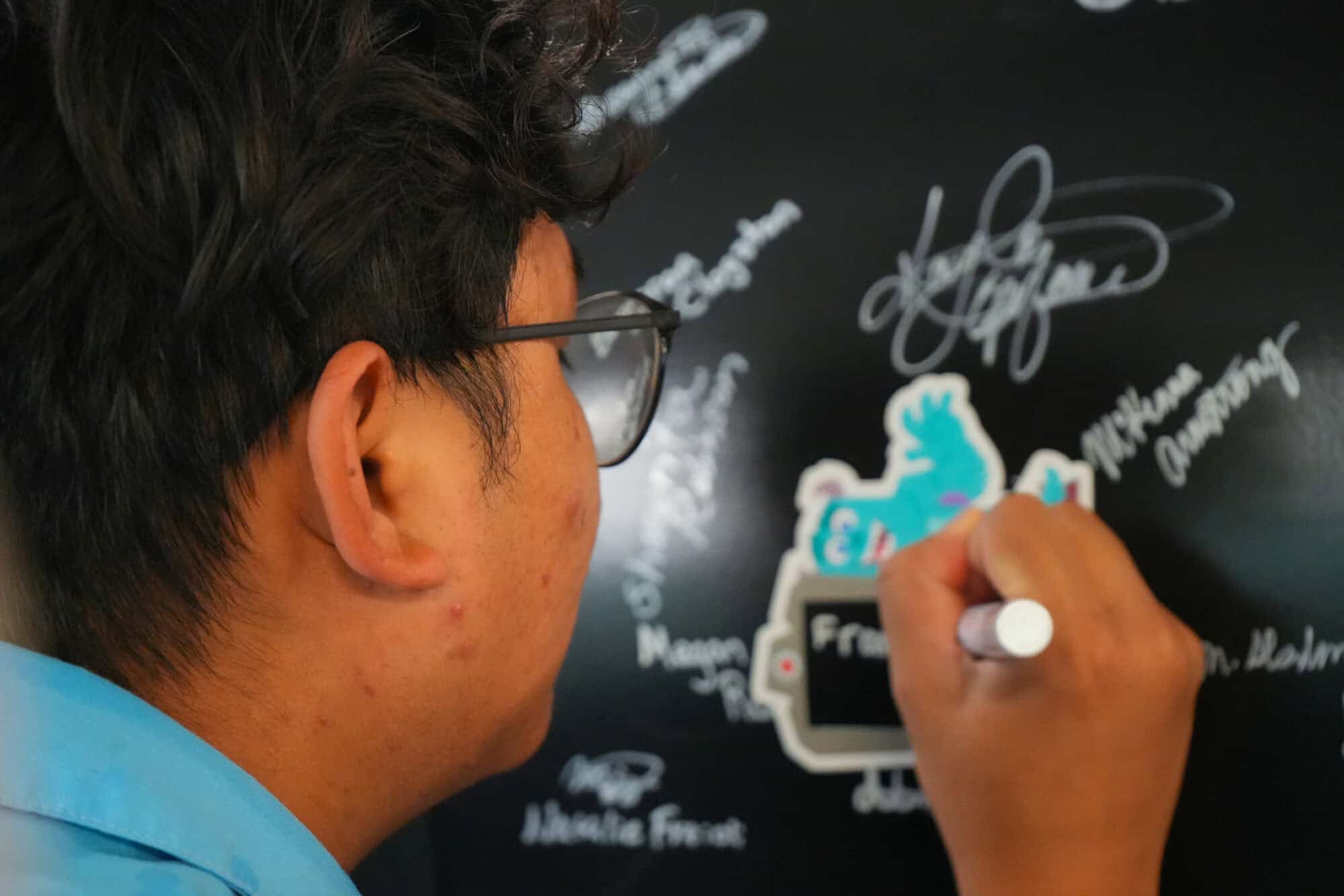 Disney theme park cast member signs a coaster beam on a black board with signatures.