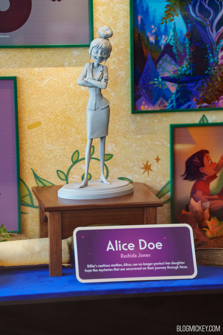 Alice Doe resin sculpture on a pedestal with colorful framed art in the background.